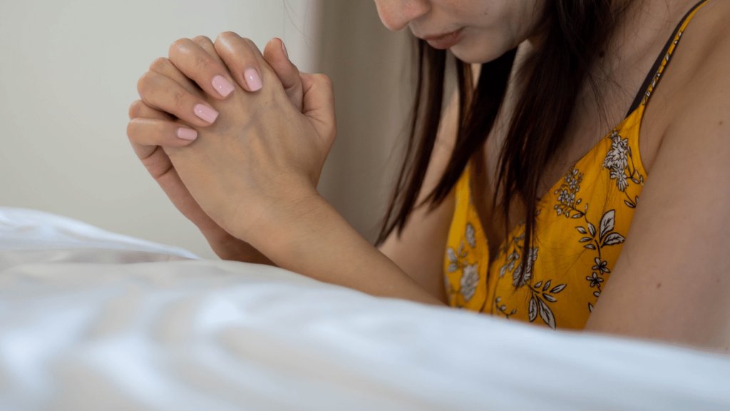 Woman in flowery shirt kneels at her bedside for sleep prayers.