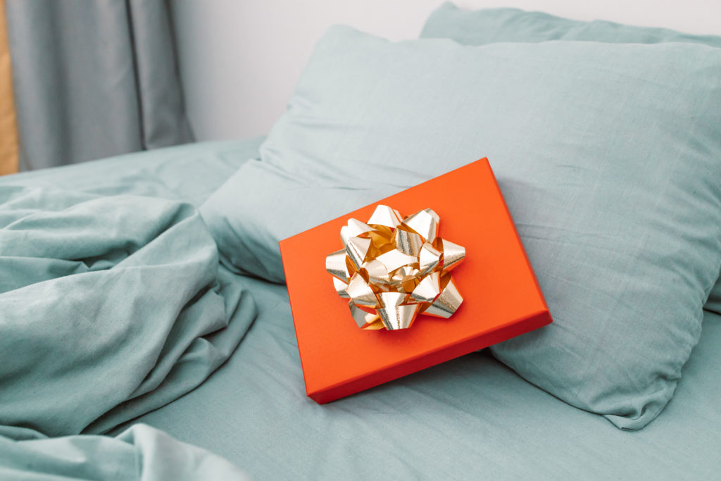 A red gift with a large gold bow leans against a pillow on a bed reminding us that sleep is a gift from God and He can help you sleep.