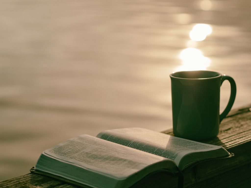 An open Bible sits on a table next to a cup of coffee next to a lake where we learn what is guided meditation.
