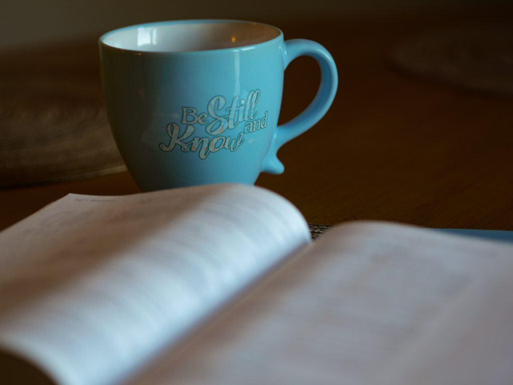 A teal blue coffee mug with the words "Be still and know" on it sits beside an open Bible where a morning prayer for peace can start your day off right.
