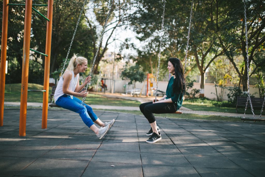A blonde woman in white sleeveless shirt and jeans, and a black-haired woman in dark pants and green shirt sit on swings at a park as they pray a morning prayer about friendship.