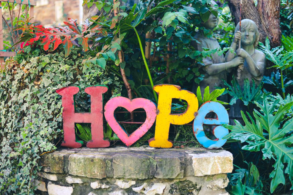 The word "hope" in colorful letters with a heart as the O sits on a stone pedestal in a garden when one can pray a morning prayer for hope and remember that God is love.