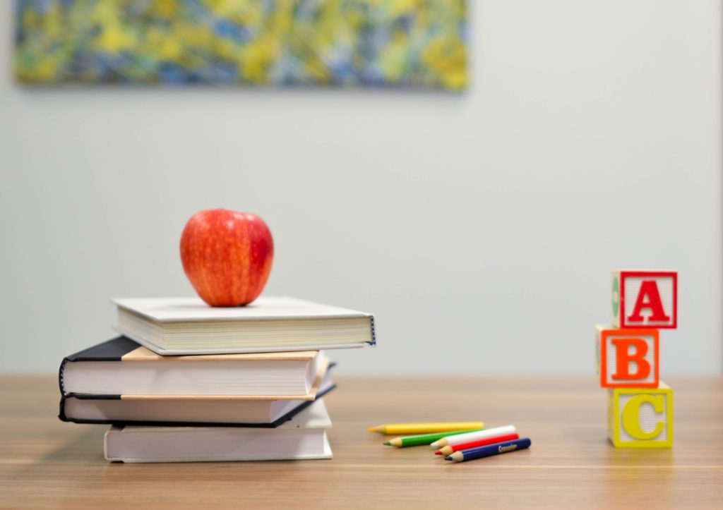 A red apple sits atop a small stack of school books on a desk with colored pencils and ABC blocks where a one-minute prayer for a new school year is prayed.