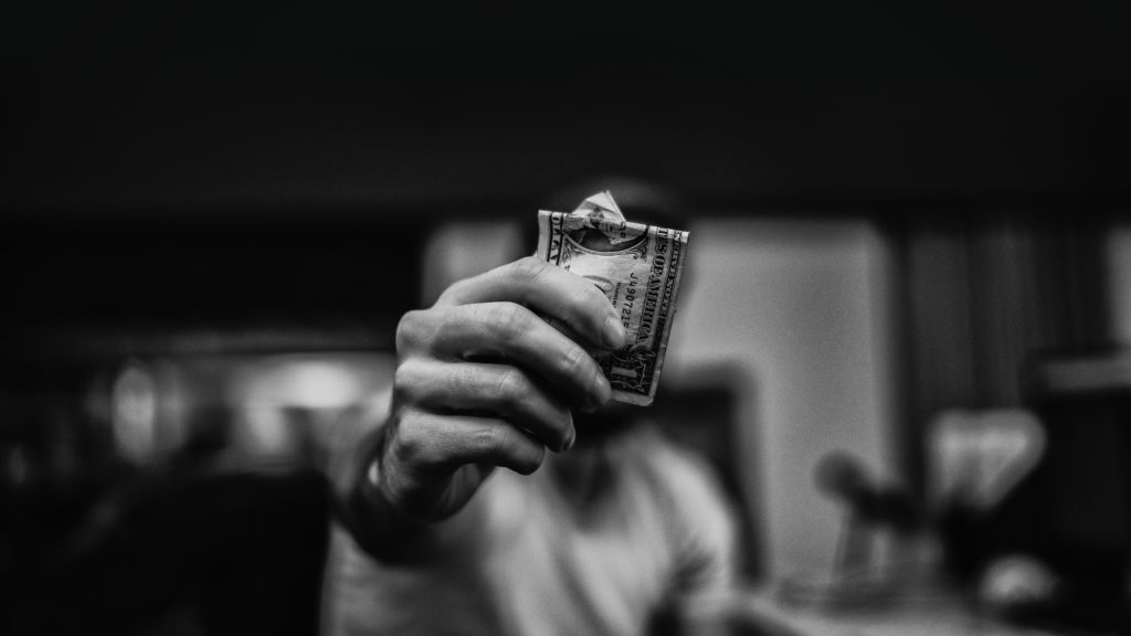 A man blurred in the background holds out a dollar bill knowing that giving can ease financial stress.