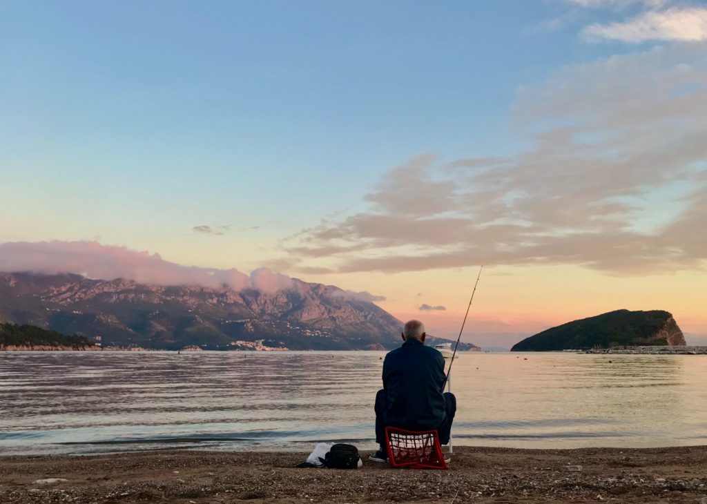 An elderly man sit on a red seat on the shore of a lake, quietly fishing after praying a morning prayer for patience.