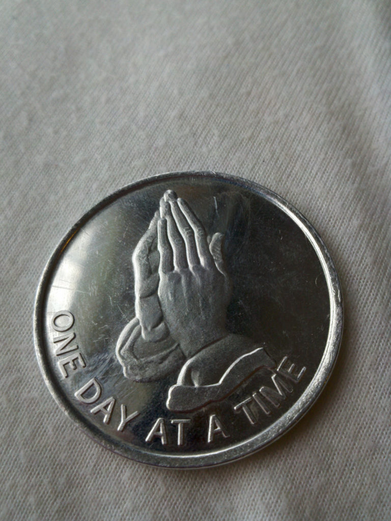 praying hands on a coin that reads "one day at a time" remind us to pray a short prayer for National Sober Day.