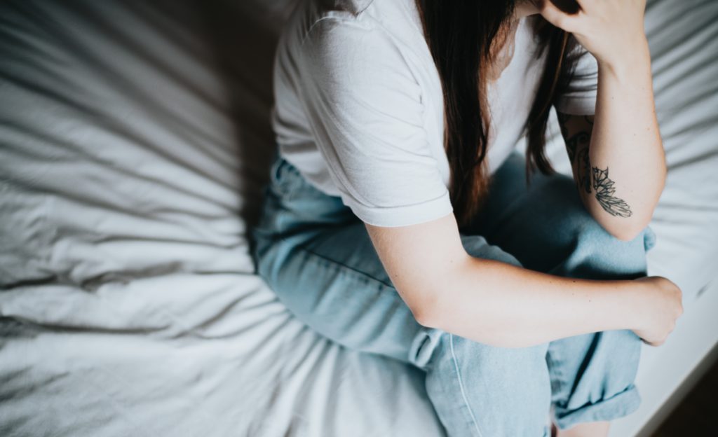 A woman in a white T-shirt and jeans sits on the edge of her bed praying a short prayer for social anxiety.