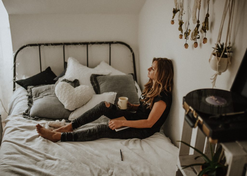 Woman leaning against a wall while sitting on a bed holding a mug prays a morning prayer to be brave.