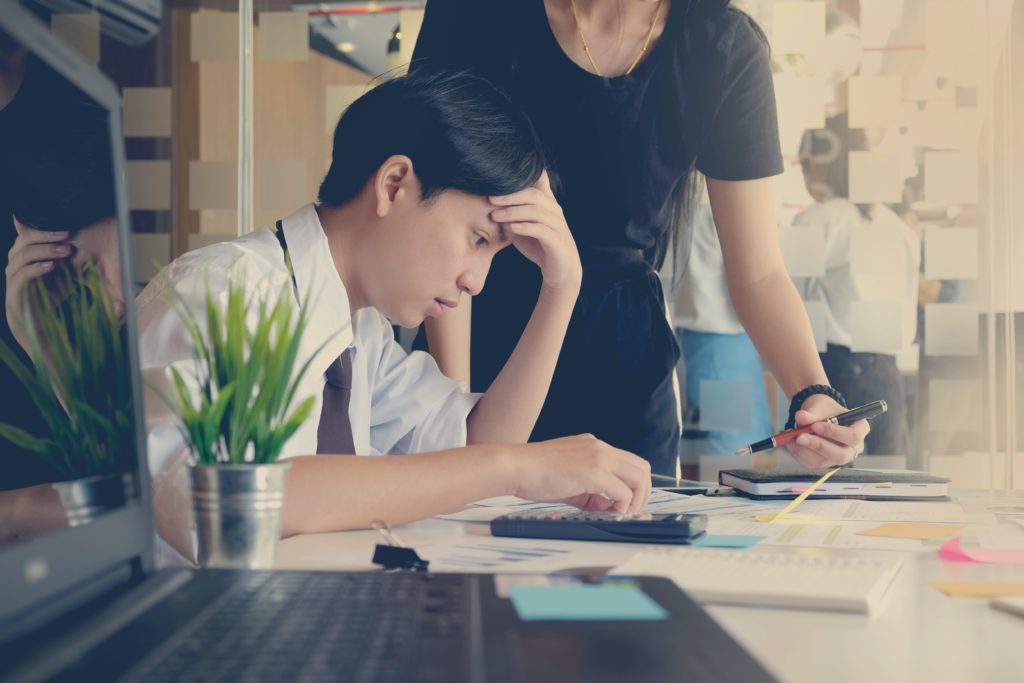 A young man in need of a short prayer for work-related stress sits at his desk in a white shirt sits while his boss points out something with a pen.  