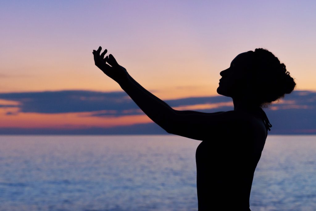 A woman in silhouette by the ocean raises her hand as she prays a prayer for mental health.