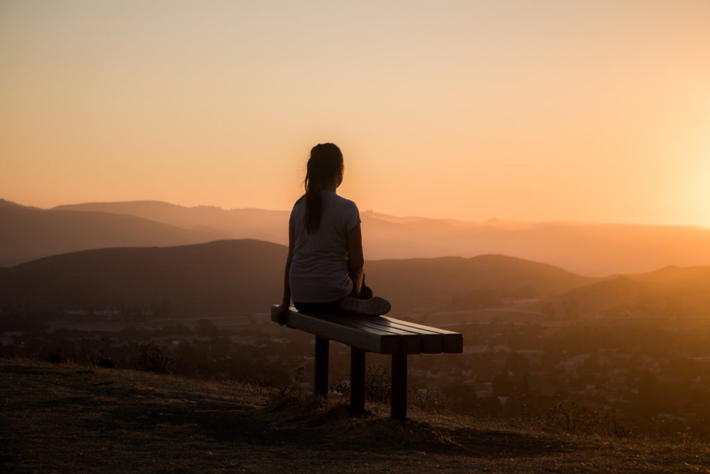 A woman sits on a bench above the hills praying a morning prayer for calm.