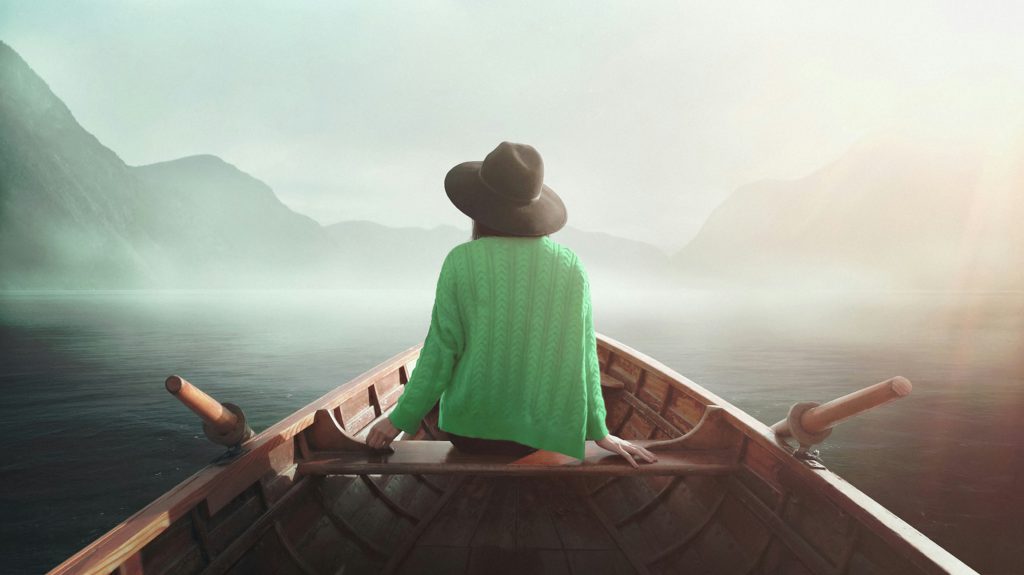 A woman in a brown hat and green sweater sits in a rowboat on a misty lake enjoying solitude.