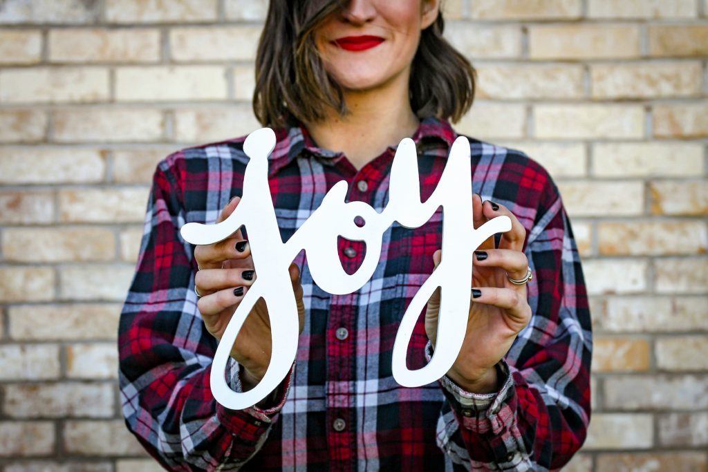 A woman in a checkered shirt holds a sign with the letters J-O-Y to aid people in finding joy.