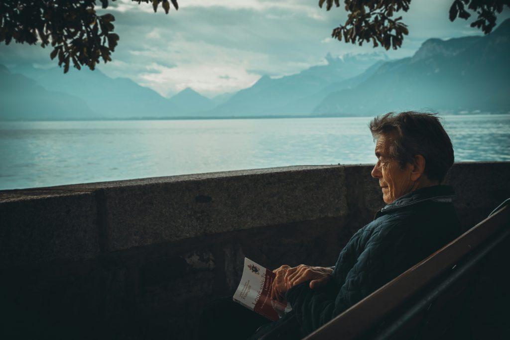An elderly man sits on a porch gazing at a mountain surrounded lake knowing that he can use Bible verses for better sleep.