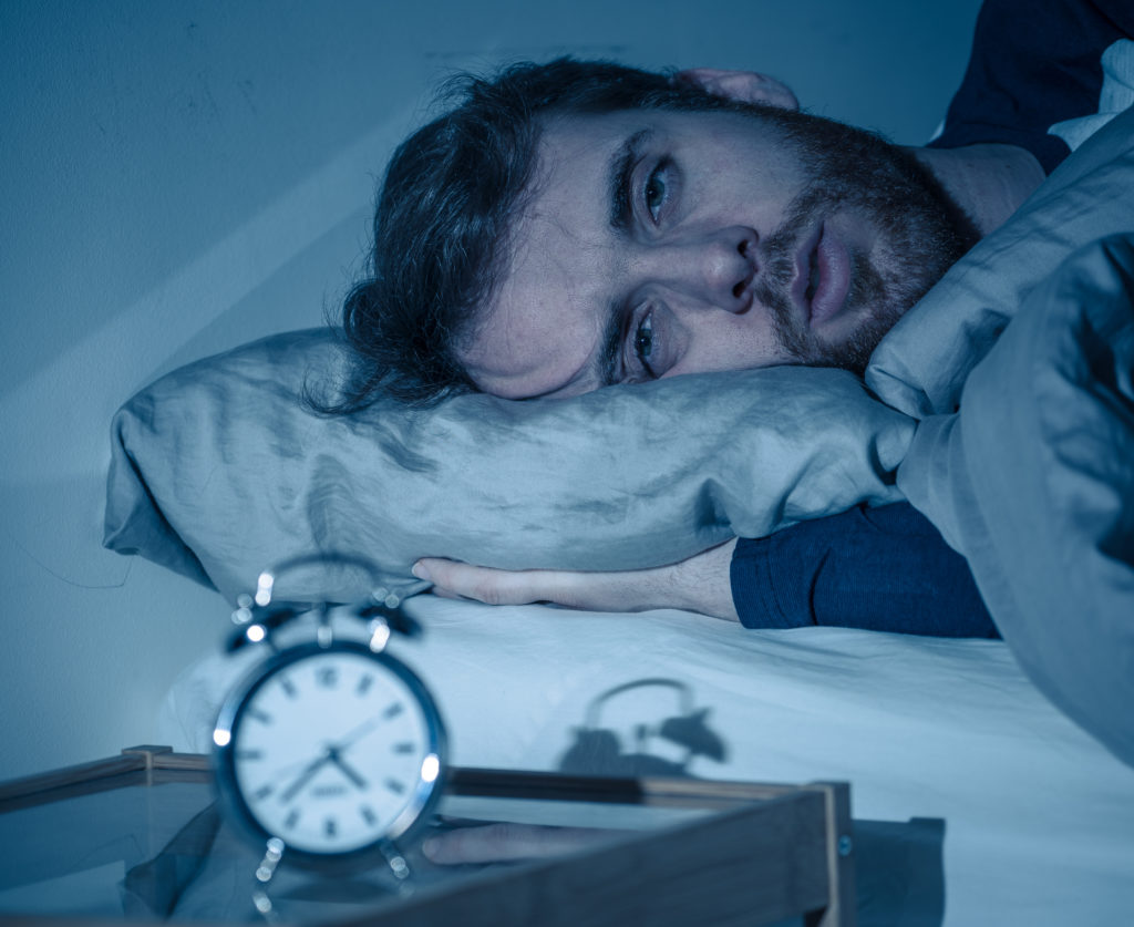 Sleepless and desperate young caucasian man awake at night not able to sleep, feeling frustrated and worried looking at clock suffering from insomnia could benefit from Christian meditation and Scripture before bed.