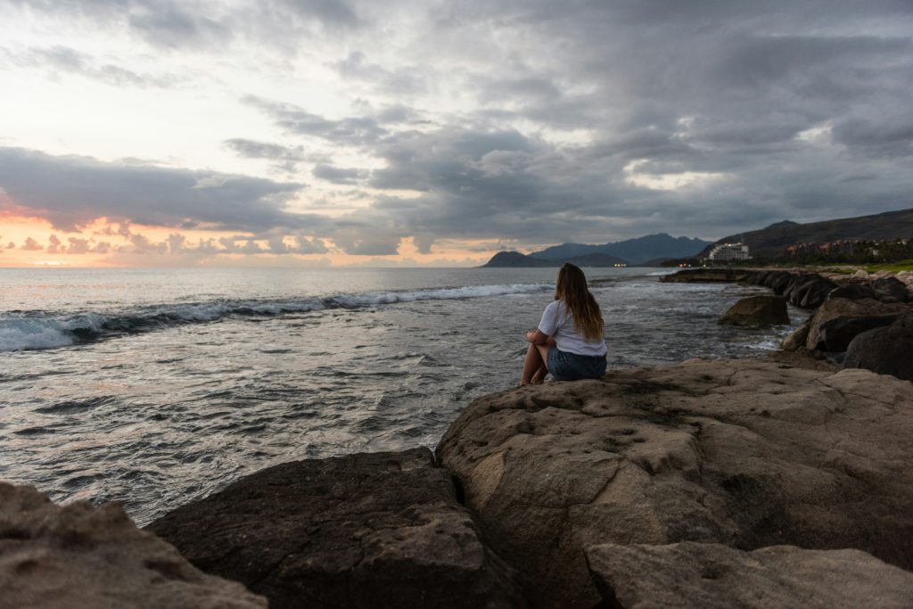 A young woman in a white shirt sits on rocks on the shore practicing Christian meditation for a good night's sleep.