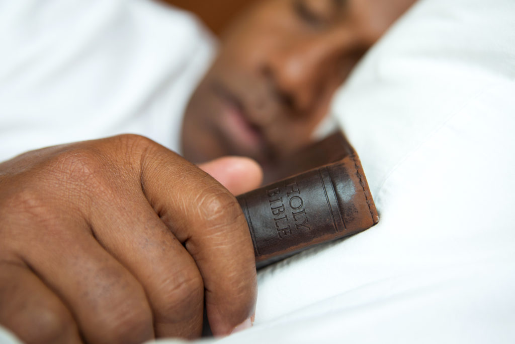 A Black man in a white T-shirt sleeps holding his Bible as a reminder to incorporate gratitude and Scripture into his sleep routine.