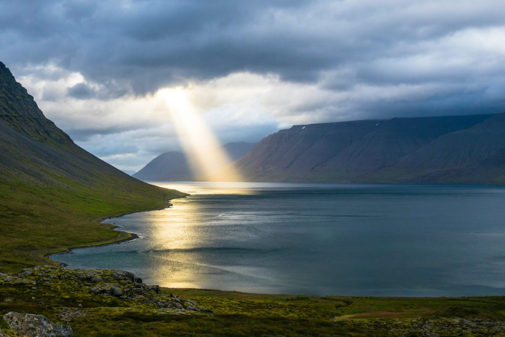God's power is displayed with a burst of sunlight through black clouds on a lake surrounded by mountains.