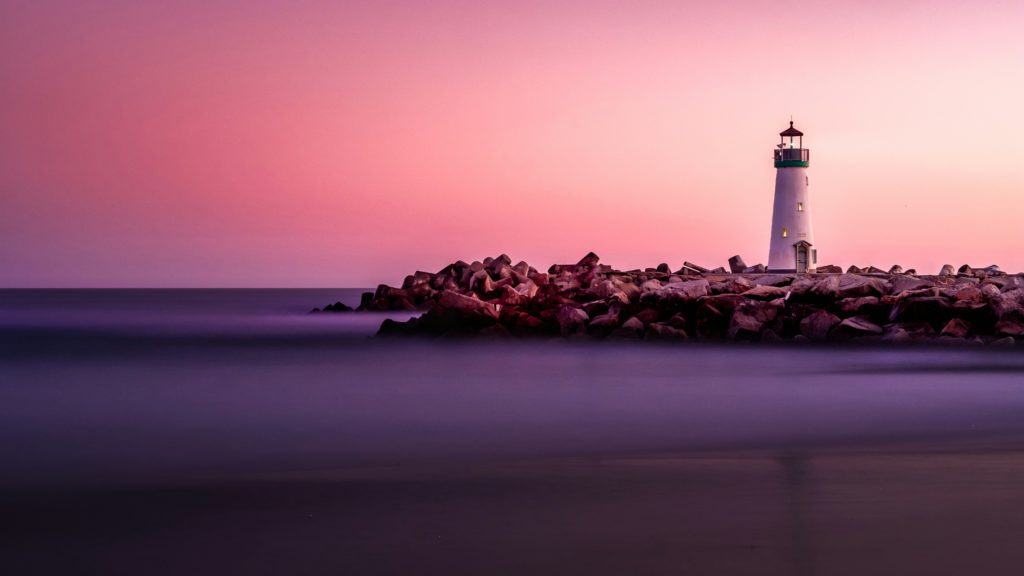 A lighthouse stands on a rocky outcropping during a brilliant pink sunrise guiding you on your quest for authenticity.