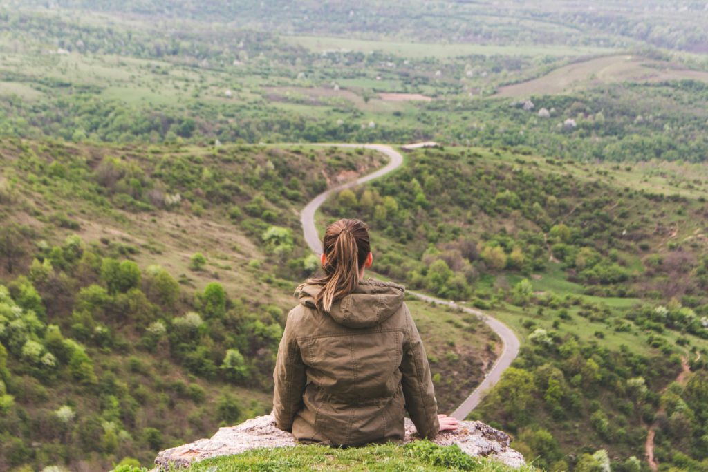 A woman sits on the edge of a rock overlooking a winding road through a grassy valley pondering the pathway to becoming unoffendable.
