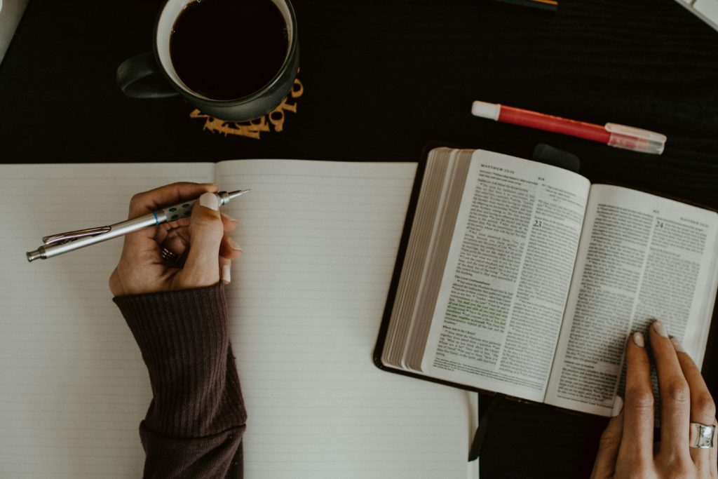 A woman's hands hold a pen over a blank notebook page along with an open Bible as she works on discerning truth.