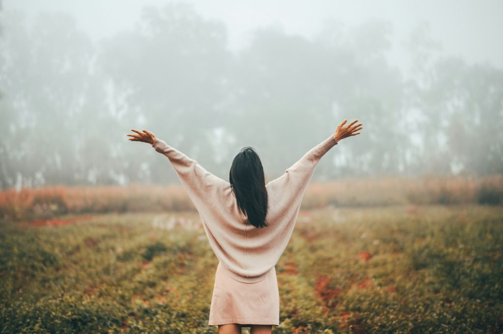 Aa woman in a light pink sweater and skirt faces a misty woods with her arms upraised learning the art of letting go.