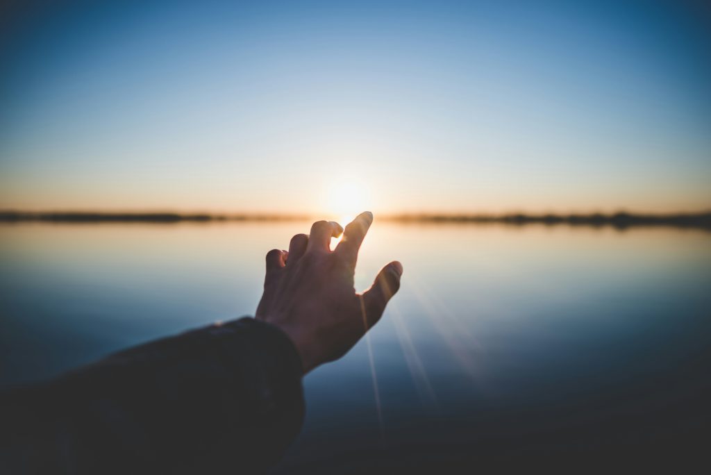 A hand reaching over the water at sunset shows God's power to heal from far off.