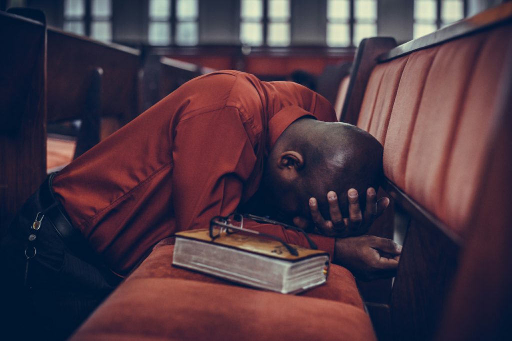 A Black man in an orange shirt kneels in front of a church pew with a Bible by his side praying fervently for God's power to heal him.