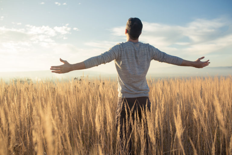 Young caucasian man standing in the middle of a prairie with his arms outstretched, enjoying a beautiful sunny day in the nature as he moves from resistance to surrender to God's will.