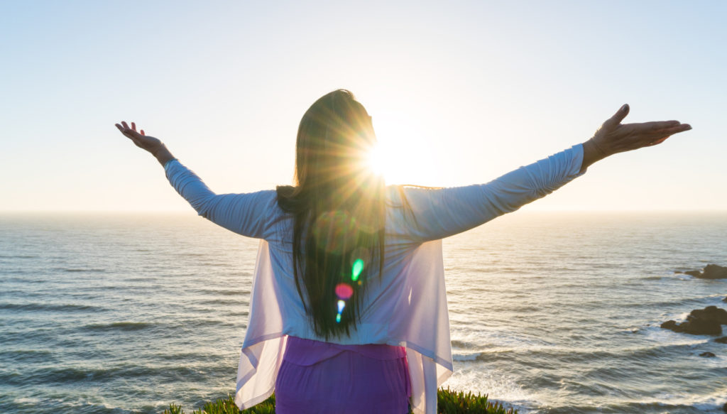 Woman with hands uplifted in worship and adoration against the warm rays of the setting sun overlooking the Pacific Ocean understands the wonder of surrendering your plans to God.
