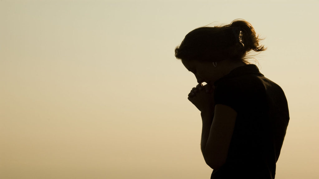 A woman prays in silhouette at sunset wondering if it's wrong to question God.