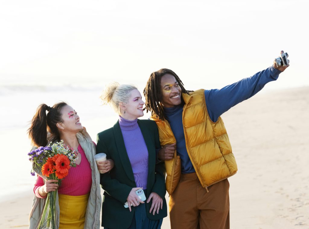 A Black man, a white woman and an Asian woman holding flowers takea. selfie at the beach, enjoying a harmonious life together.