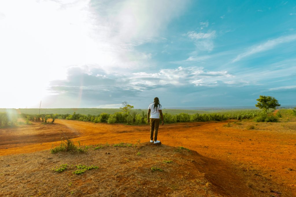 A man in a white T-shirt stands on a dirt mound overlooking dry bushes battling doubts.