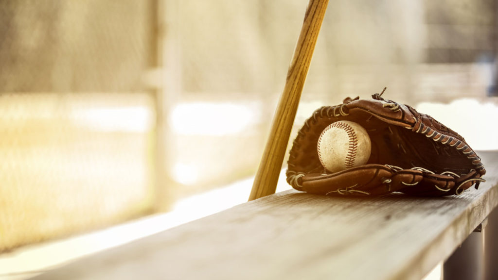 A bat, ball and glove sitting on a wooden bench beside a baseball diamond illustrate what it feels like to be benched.