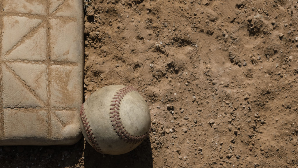 A baseball sits next to a base on the infield dirt with the symbol of a cross reminding us that building belief is a lot like baseball.