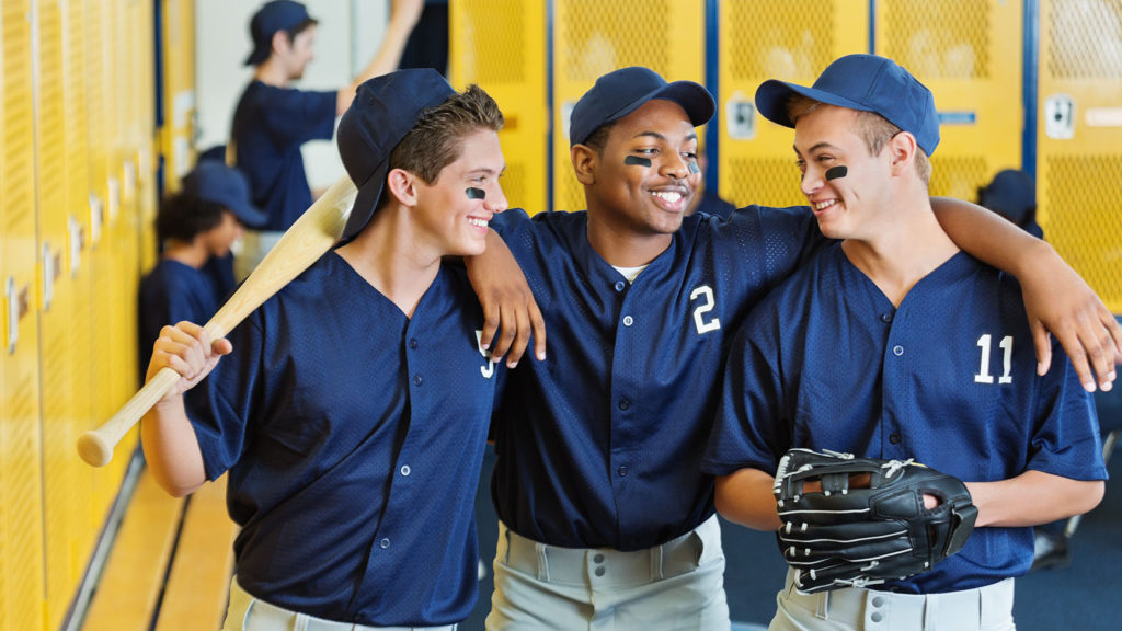 Three members of a baseball team in a locker room wearing navy blue jerseys and holding  baseball equipment are building belief as they play the game.