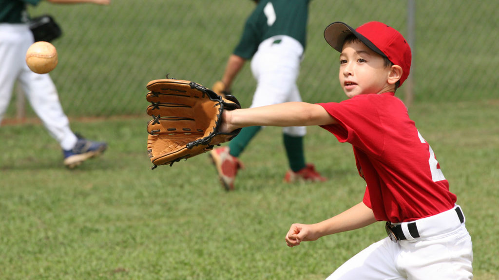 A young Asian Little League baseball player wearing a red hat, red team shirt, and white baseball pants eagerly catches a thrown ball like he's catching hope.