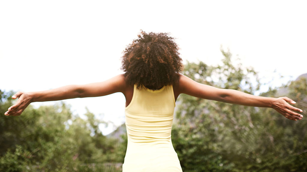 A young Black woman stands with her arms wide open as she rests in the knowledge that she is cherished by the Creator.