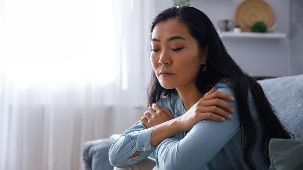 A distraught young woman sitting on the sofa, hugging herself and looking down while struggling with understanding her place in the divine tapestry.