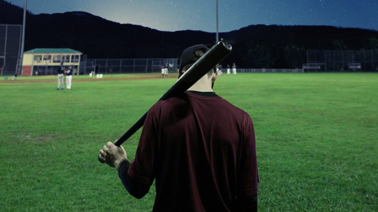 A baseball player stands in the outfield with his bat on his shoulder, tired during a game that went into extra innings.
