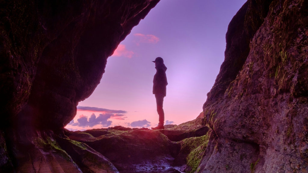 A woman stands in silhouette at the mouth of a large cave as she loos out at a purple and pink sunrise giving thanks to her faithful God.