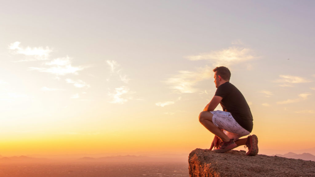 A man squats on a cliff edge at sunset as he gets his game on for God.