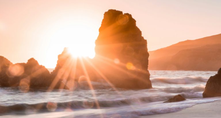 A setting sun through rock formations on the beach remind us to hold on to hope.