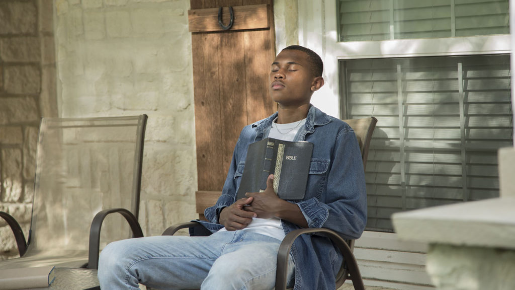 A young Black man sits on his front porch with his open Bible learning to slow down and find lasting contentment as he prays with closed eyes.