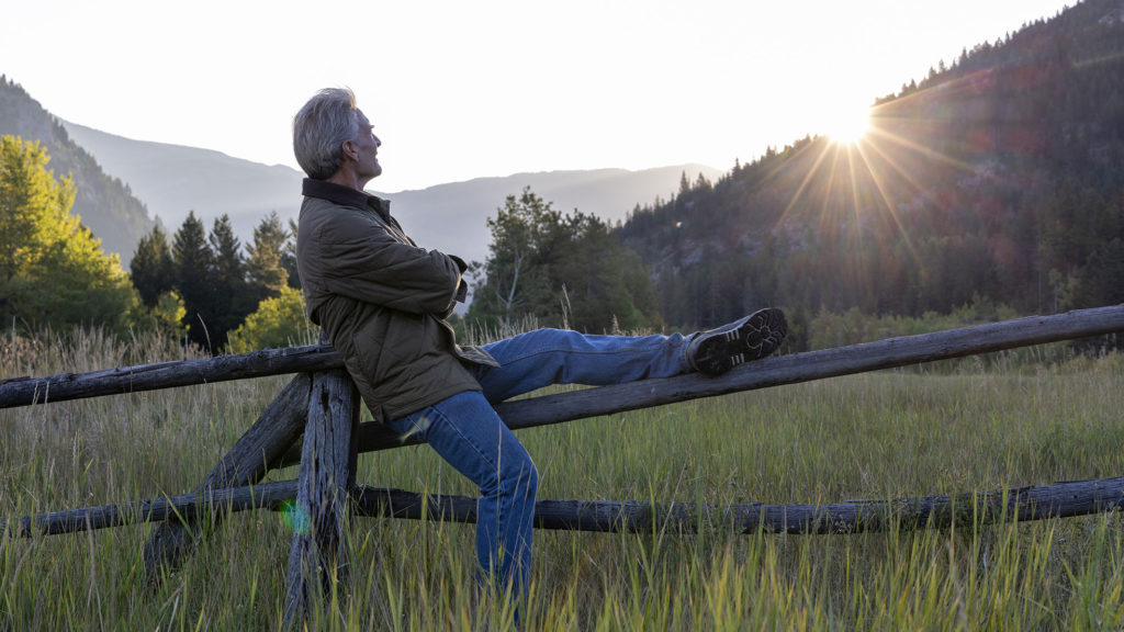 A man sits on a split rail fence watching as the sun rises over the mountains in distance, Crowsnest Pass, Alberta finally learning how to slow down.