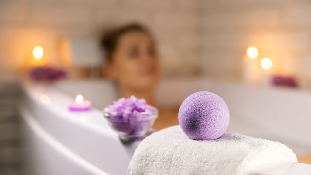A woman blurred in a tub uses a lavender bath bomb and candles to see if they help with unlocking serenity.