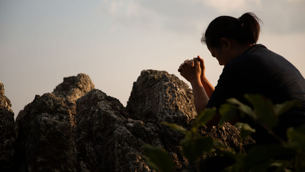 A woman prays at dusk amidst large rocks wondering if it's wrong to question God.