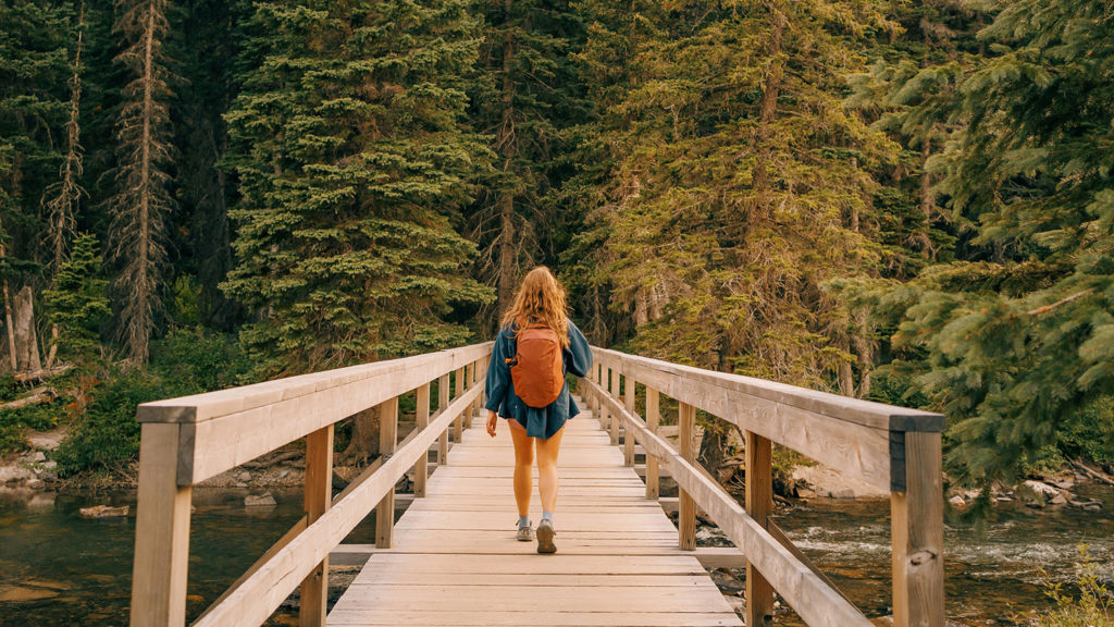 A woman wearing an orange backpack walks on a bridge in the woods as she thinks about how to let God carry it.