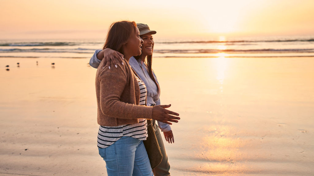 An older woman and a younger woman walk on the beach at sunset as they are growing together in their faith.