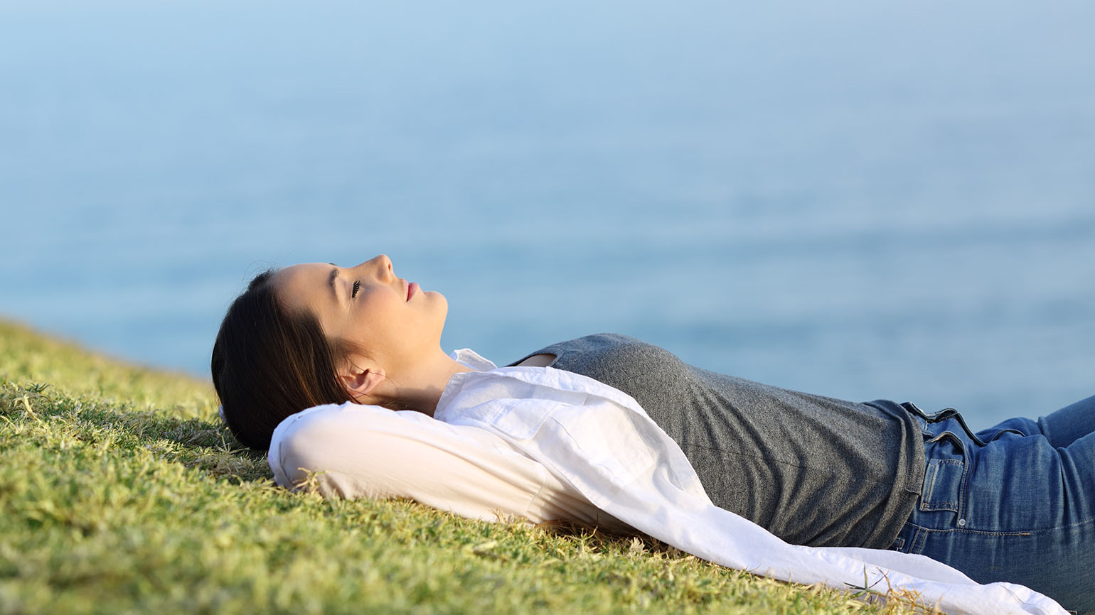 A woman in a grey T-shirt and jeans with a a white coverup sleeps on a hillside overlooking the ocean as she experiences spiritual serenity.